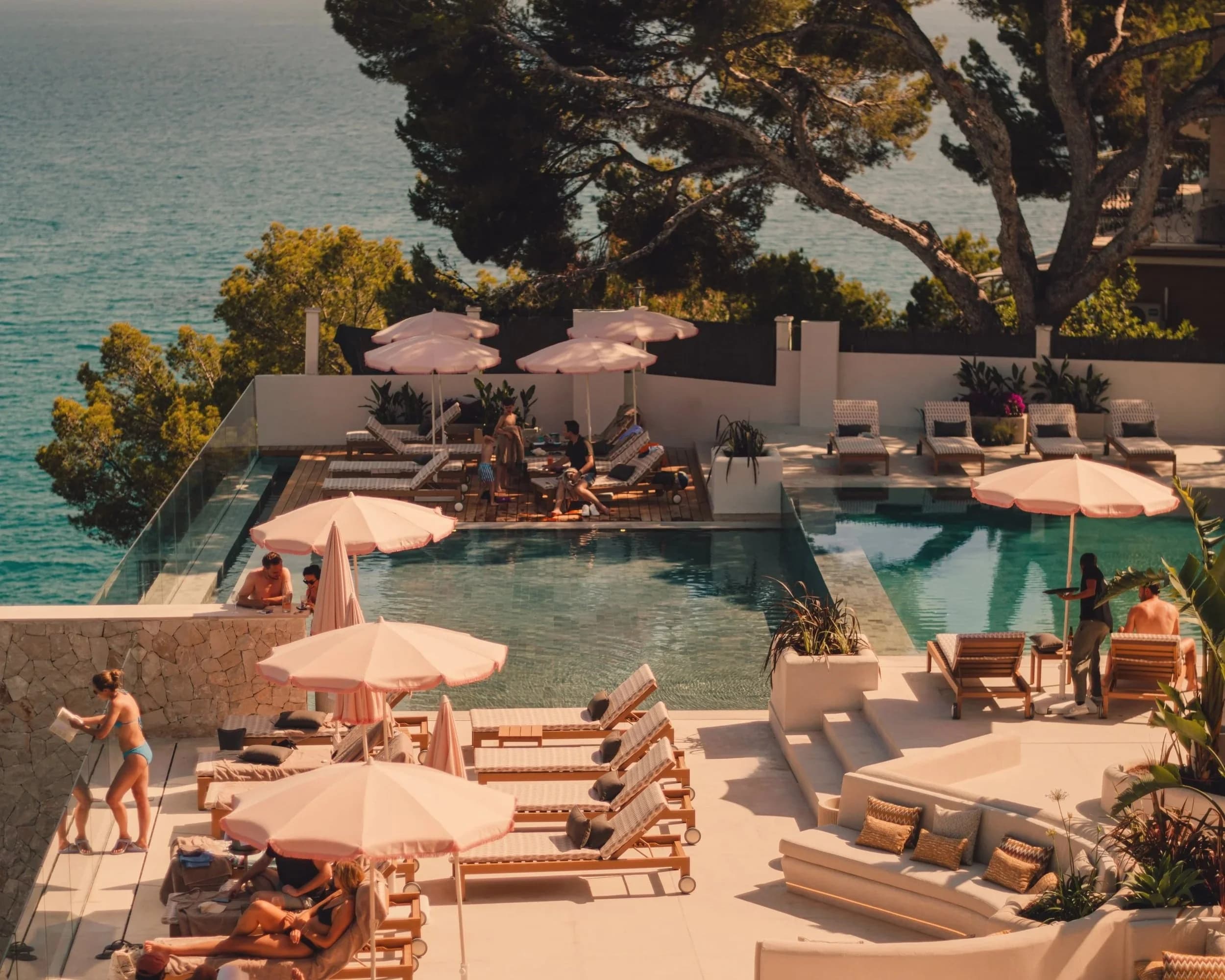 Swimming pool area at a resort with lounge chairs, umbrellas, and people relaxing and socializing near the pool, overlooking a body of water with trees in the background.
