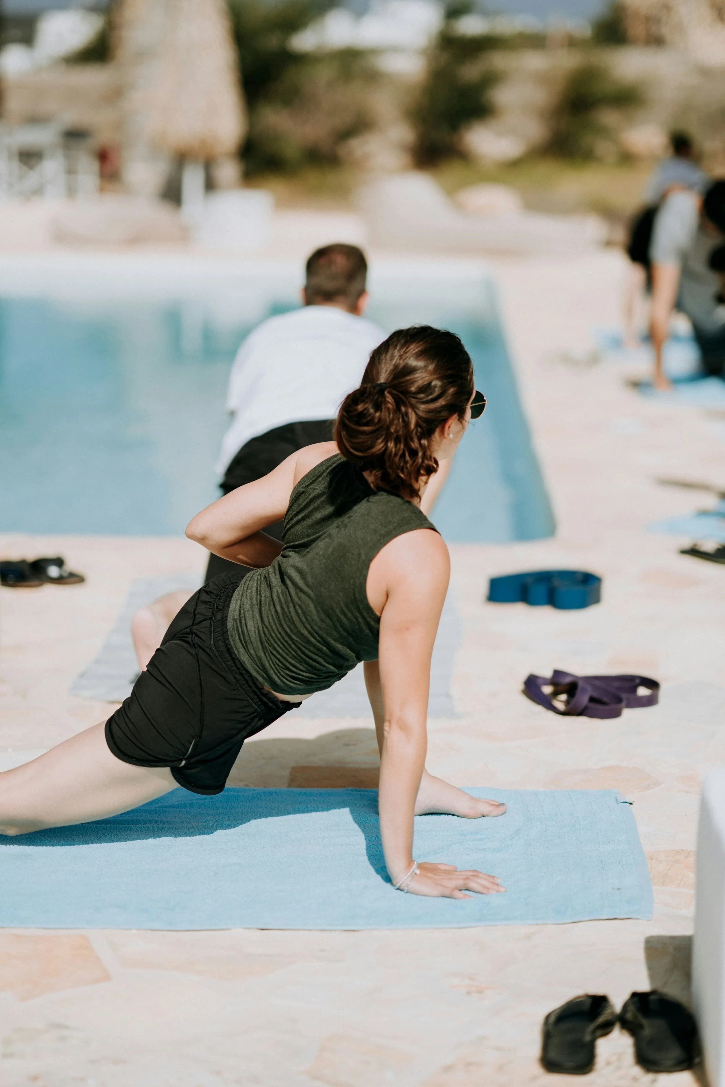 Woman doing yoga poses near a swimming pool, with other people in the background indicating a relaxing outdoor setting.