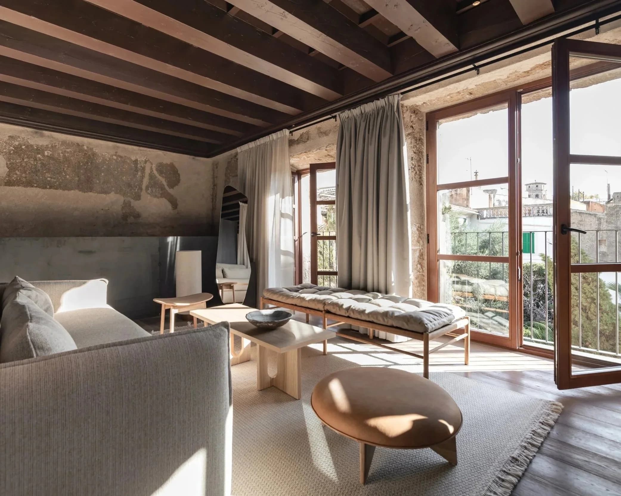 Modern living room with rustic elements, featuring a beige sofa, wooden coffee table, cushioned bench, round ottoman, and large windows with curtains, letting in natural light.