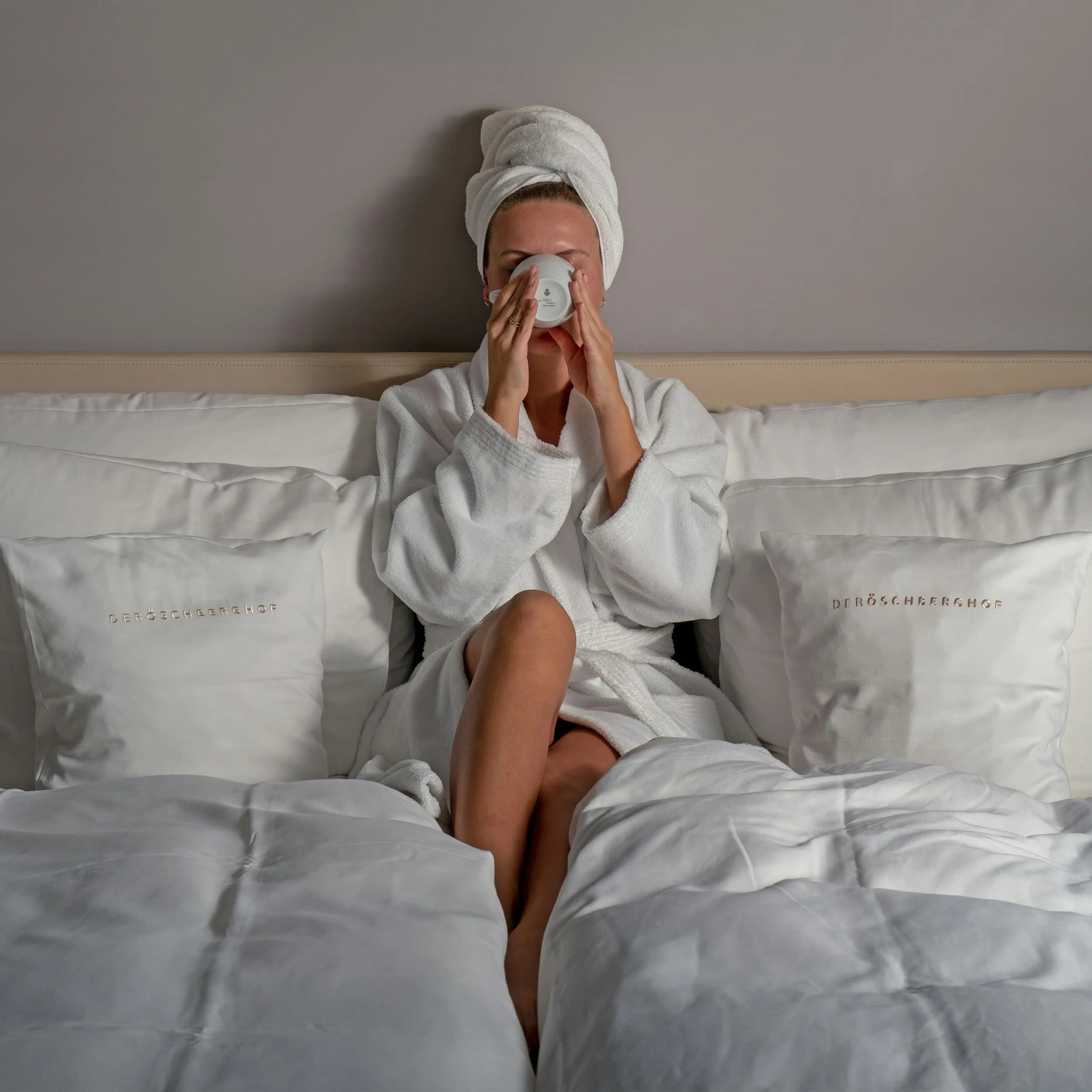 A woman in a white bathrobe and towel on her head sitting on a bed, drinking from a white mug.