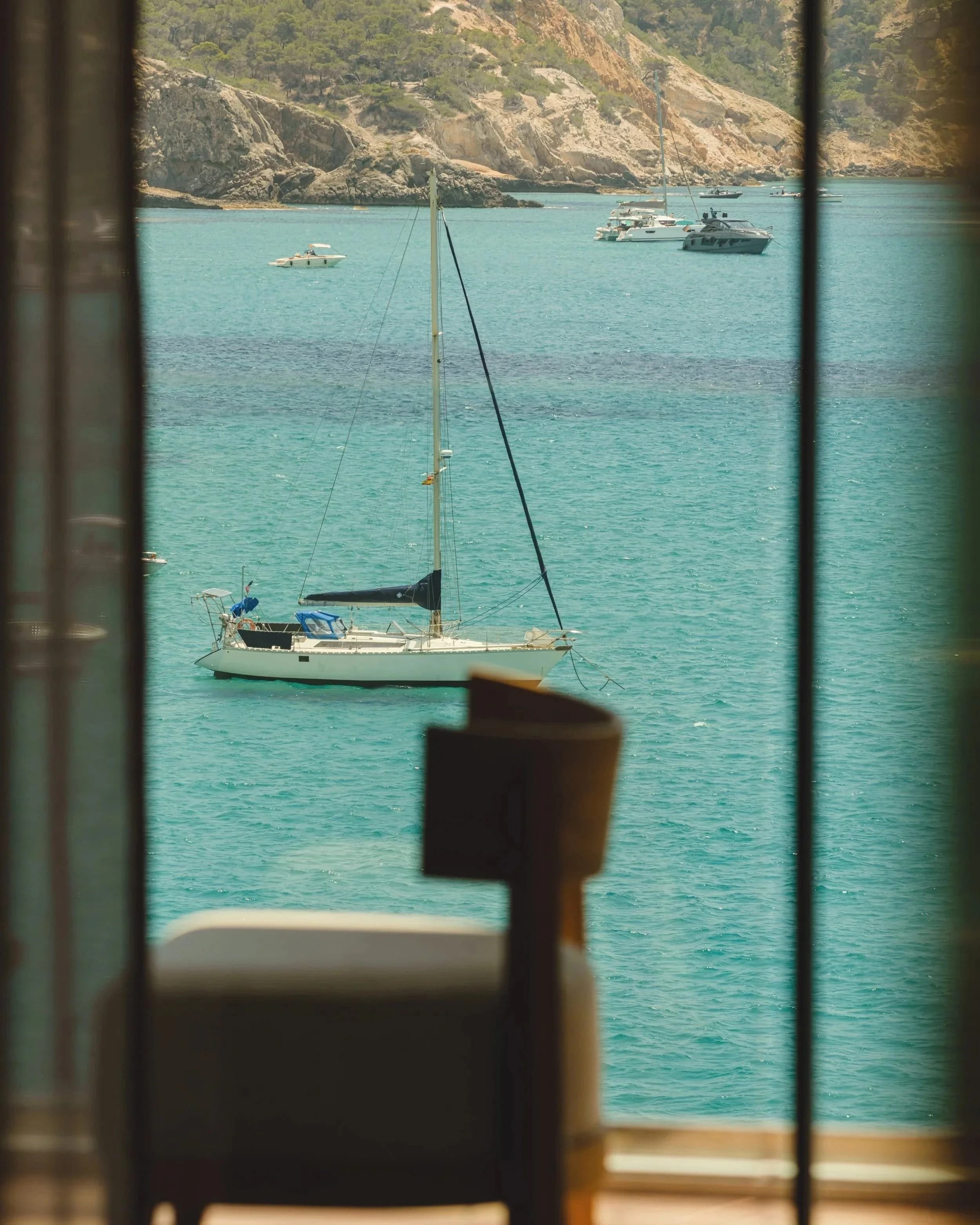 View of sailboats on calm turquoise water through window with curtains, with rocky hills in the background.