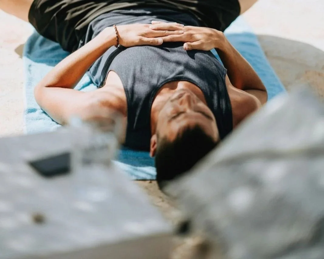 A person lies on their back on a blue mat, resting with their eyes closed, wearing a gray tank top and a bracelet on their left wrist.