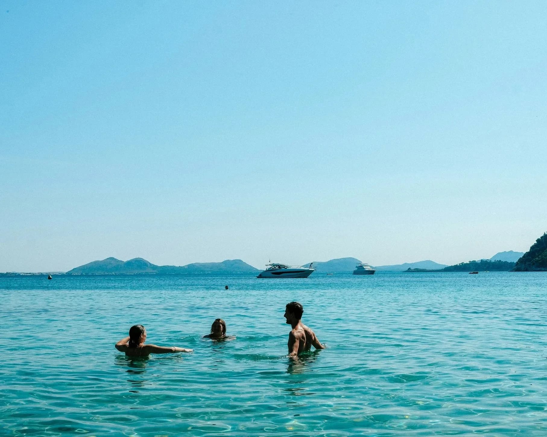 Three people, two women and one man, enjoying a swim in clear blue water with boats and islands in the background.